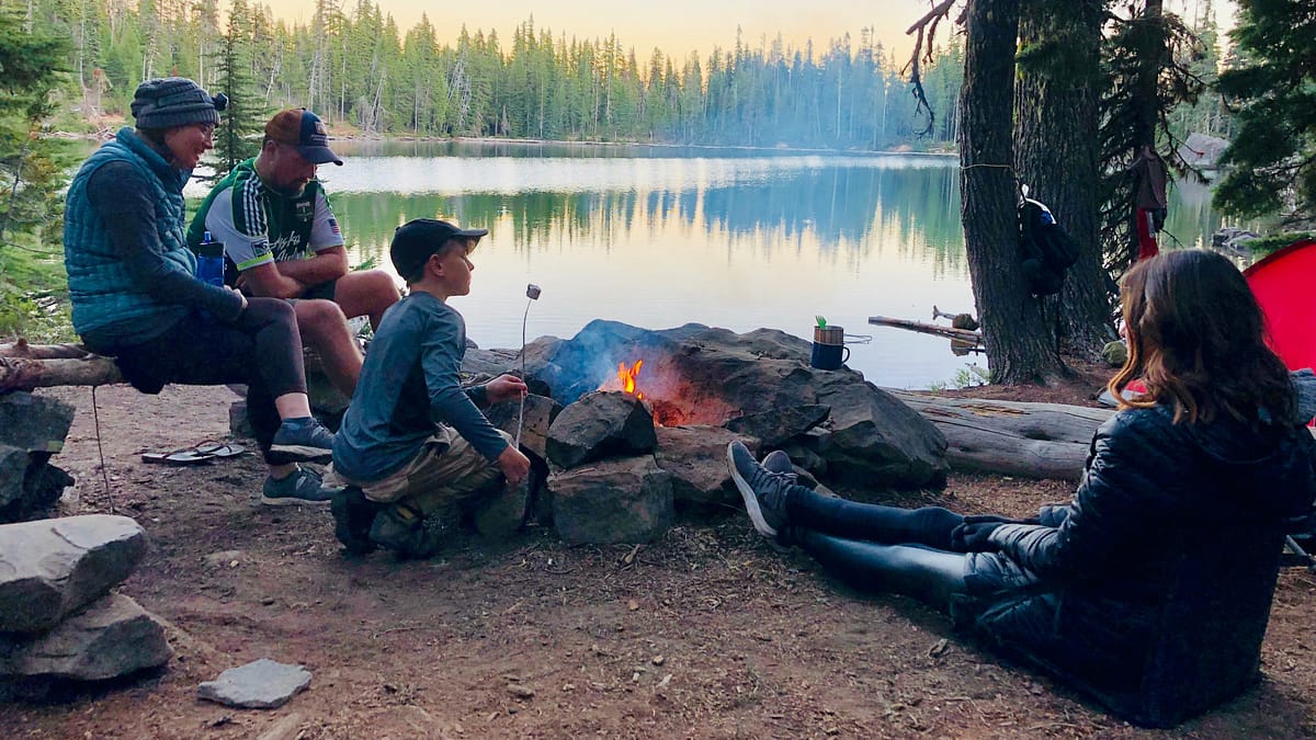 A picture of a small family sitting around the campfire in the morning by a beautiful, serene lake in a popular campground, following camping etiquette. Photo by Brooks Rice.