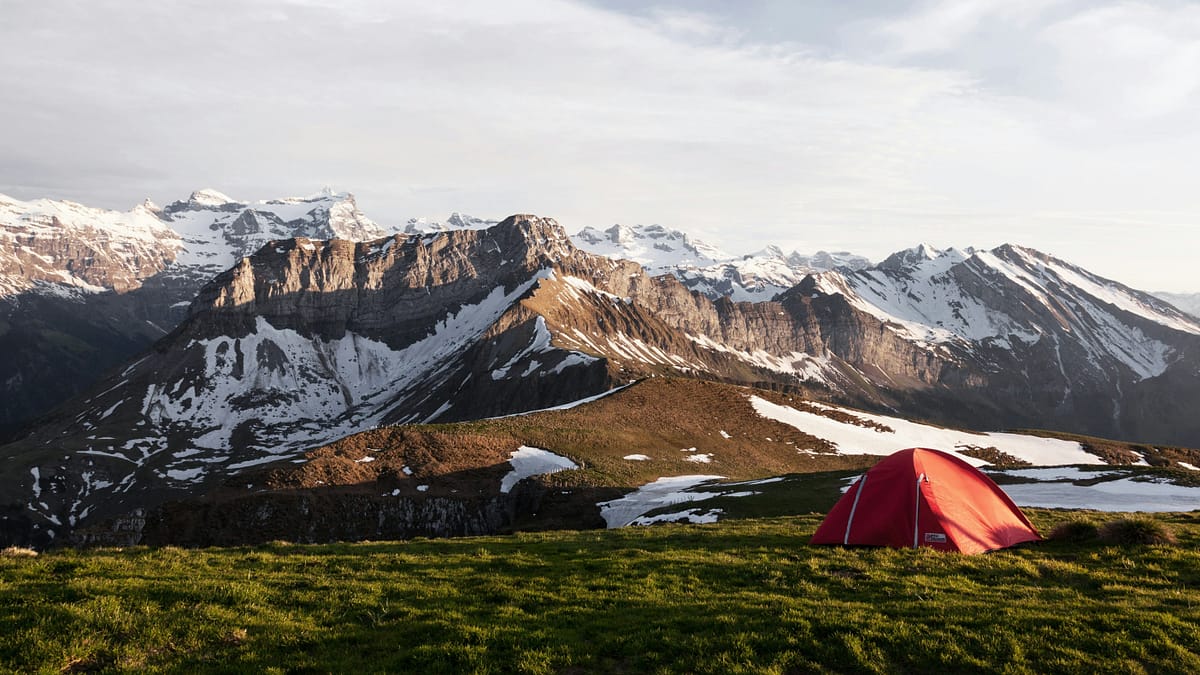 A single tent on a remote hillside overlooking a massive mountain landscape with snow-tipped mountains. Photo by Dino Reichmuth.