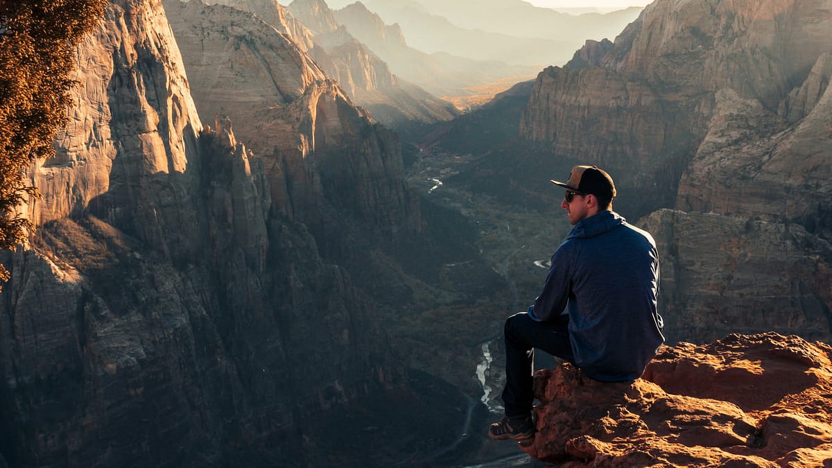 A man wearing a hat, sunglasses, a hiking coat, jeans and hiking shoes sitting on a rock overlooking a huge canyon area within Zion National Park. Photo by Jamie Fenn.