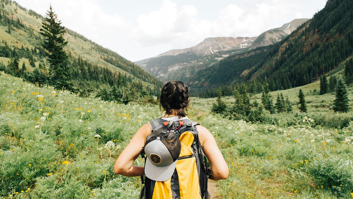 Woman hiking in the mountains, going through a valley filled with tall grass, sparse, trees, and blooming wildflowers, holding her backpack strap. Photo by Holly Mandarich.