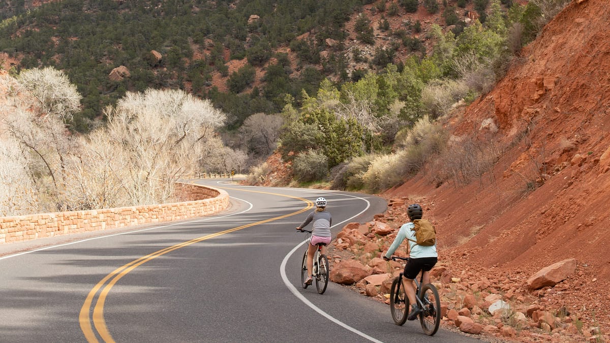 Two people, a man and a woman, mountain biking through a section of Zion National Park along the roadway. Photo by Matthew Hicks.