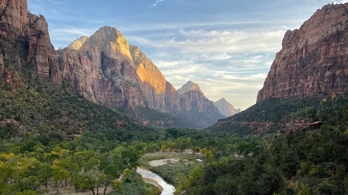 A beautiful landscape in Zion National Park on a slightly cloudy day with a small river meandering through the canyons, lush greenery on both sides - photo taken by Alex Donnachie.