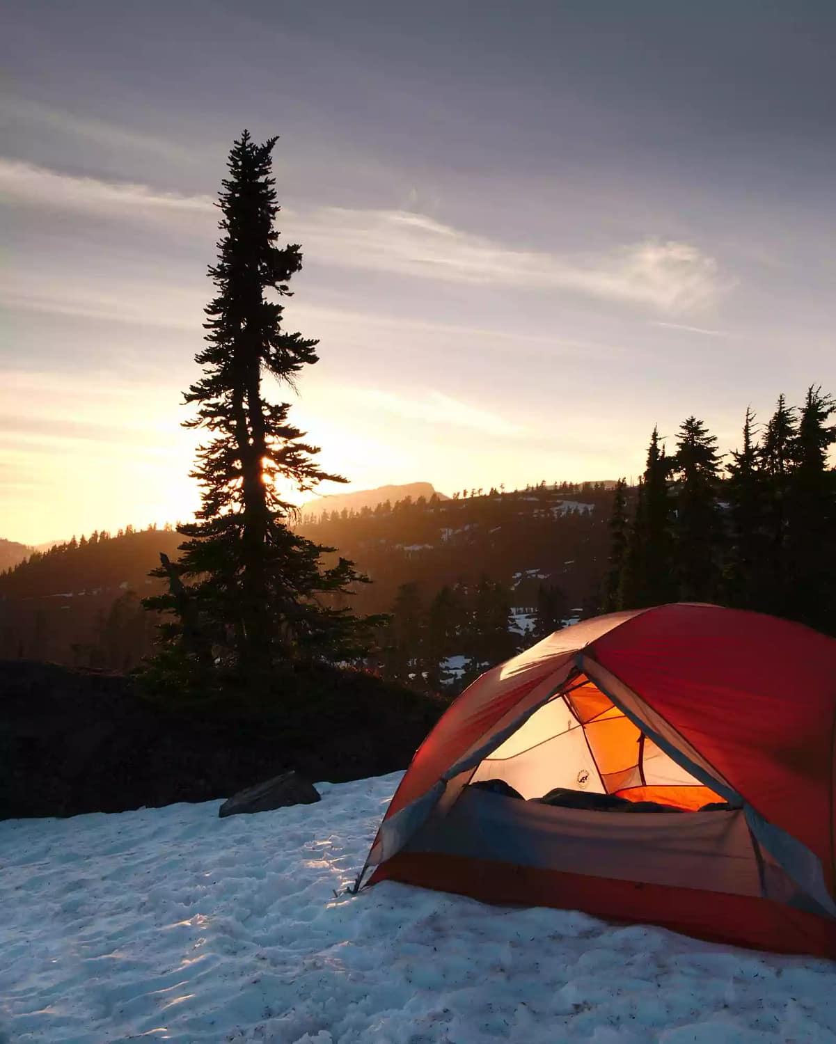 An orange dome tent pitched on a snowy spot for winter camping near a pine tree. The sun is setting behind a slope in the background.