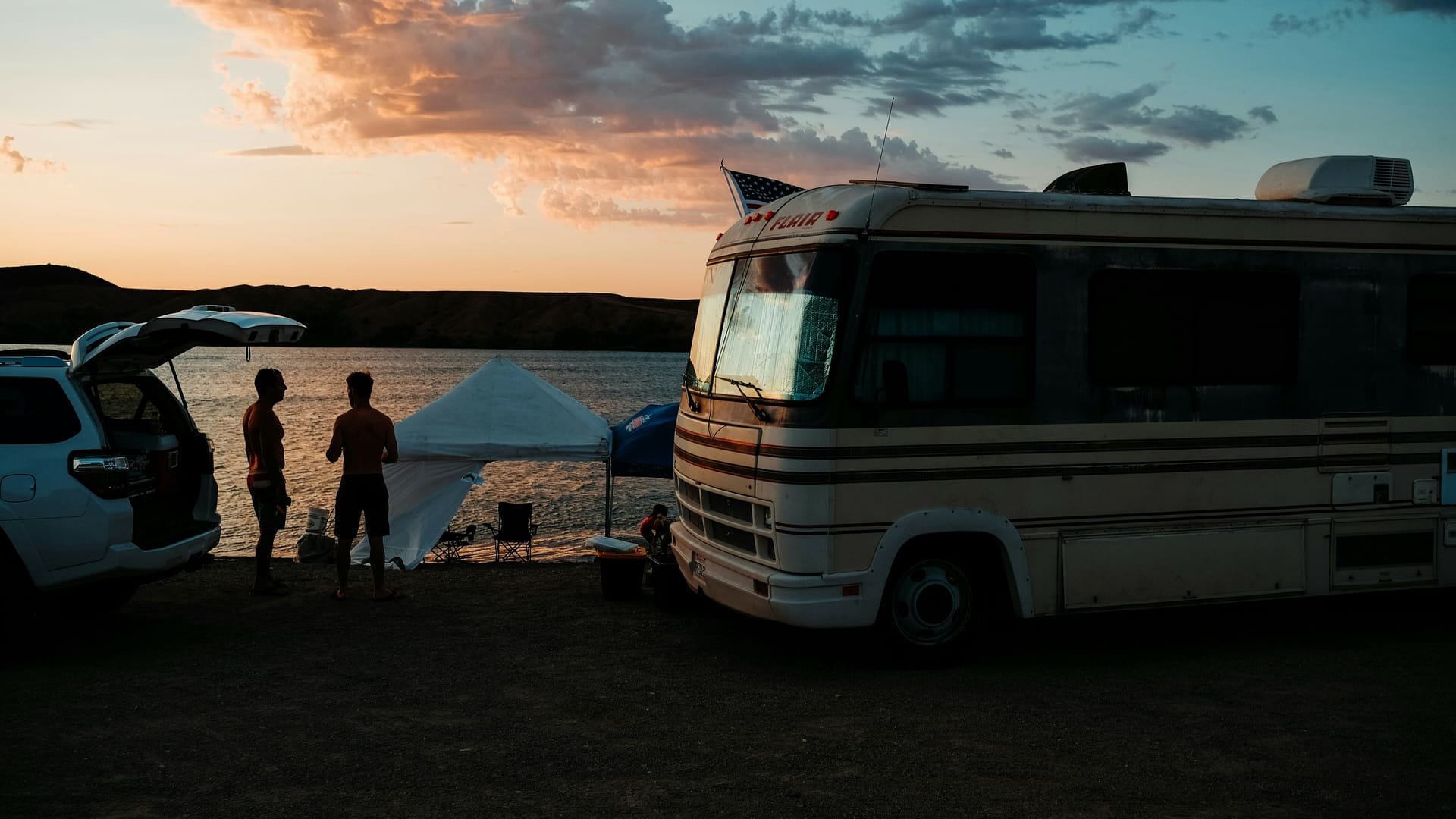 Two men standing beside a classic RV motorhome by a lake not long after sunset, by tents and camping chairs. Photo by Leon Bublitz.