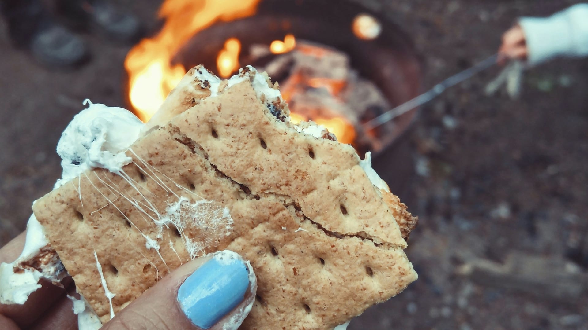 A big, thick, gooey smores being held by a woman's hand with pink fingernails, with someone cooking camping food at the fire in the background. Image by Autumn Mott.