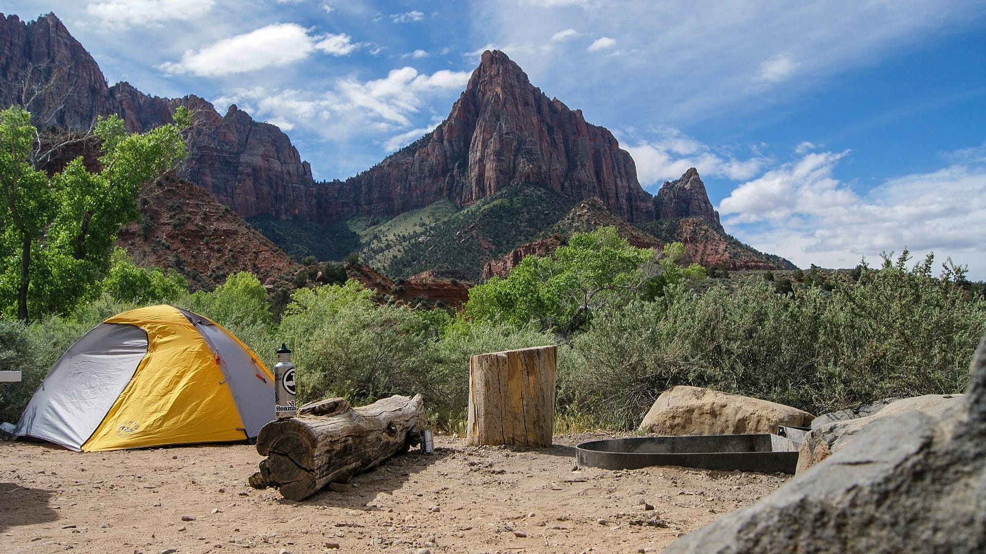 A campsite in Zion National Park with a tent, water bottle, and seating around the fire pit and large rock formations in the background.
