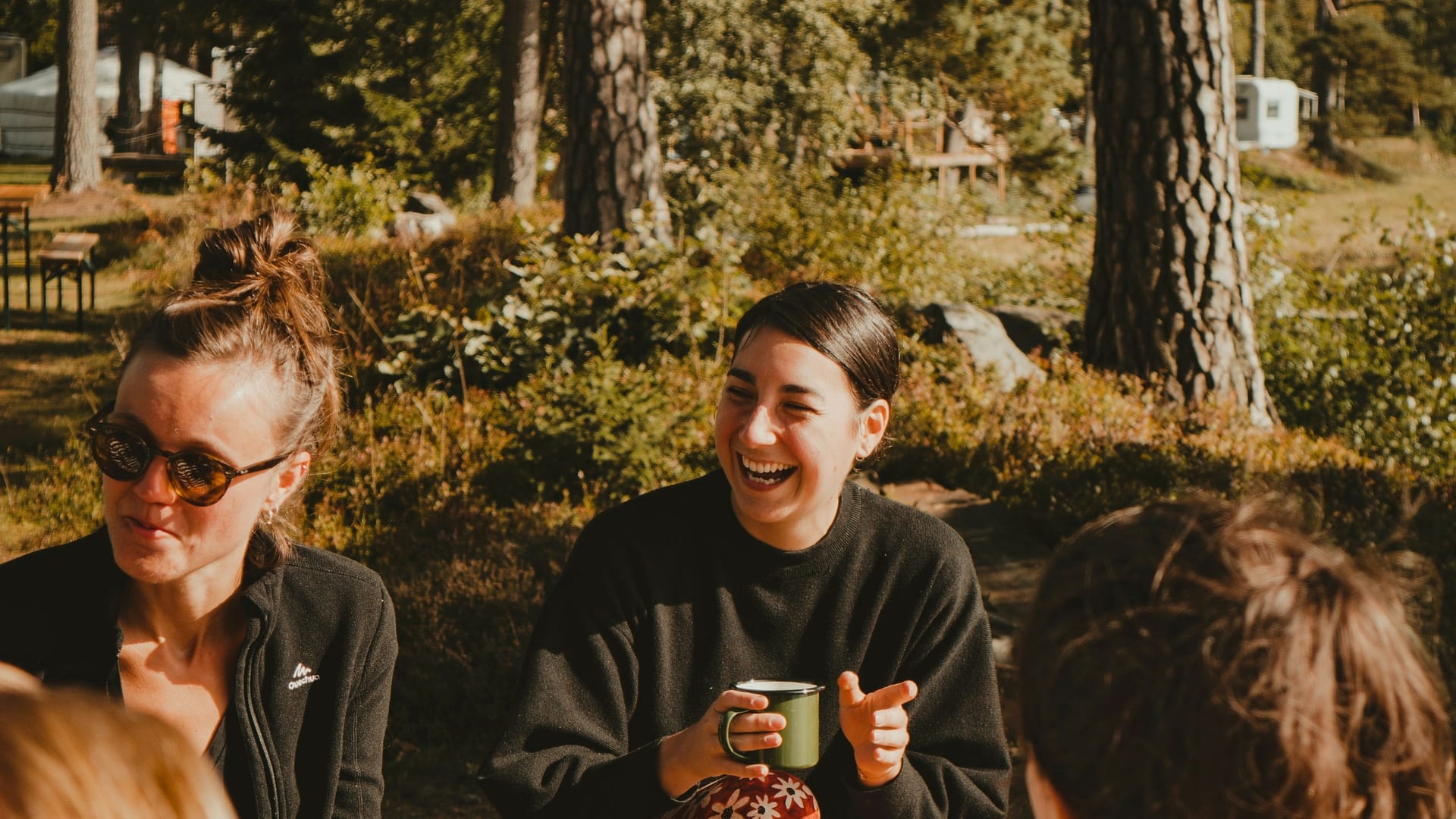 Campers at their site enjoying some outdoor hospitality on a sunny day, drinking coffee in camping mugs, smiling, and laughing with trees in the background. Photo by Tam Koppelaar.