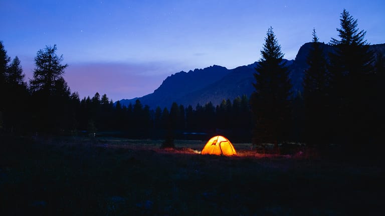 A single glowing tent camping at night alone in the wilderness with tress on both sides and mountains in the twilight distance.